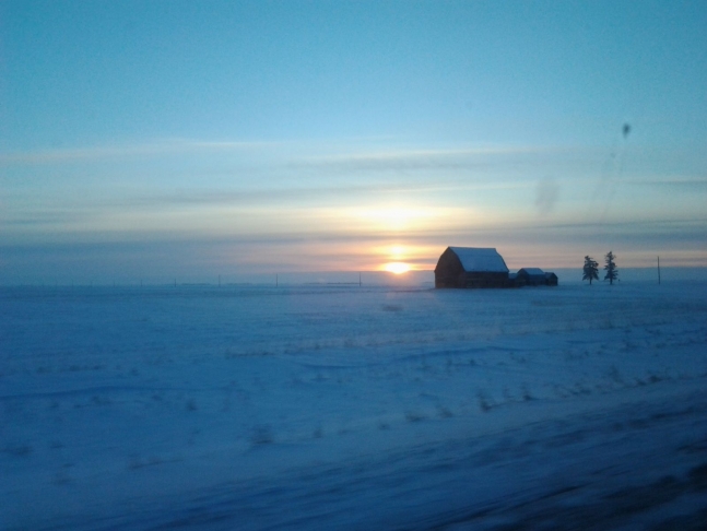  the sun rises in a cold winter sky next to a barn and outbuildings huddled on the open prairie